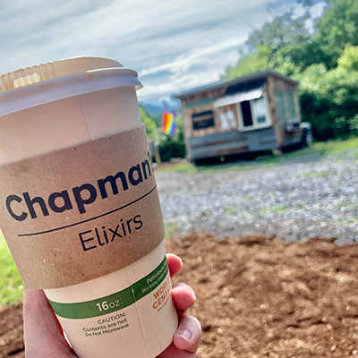A hand holding a cup of coffee with the Chapman's Elixirs store in the backgroud.